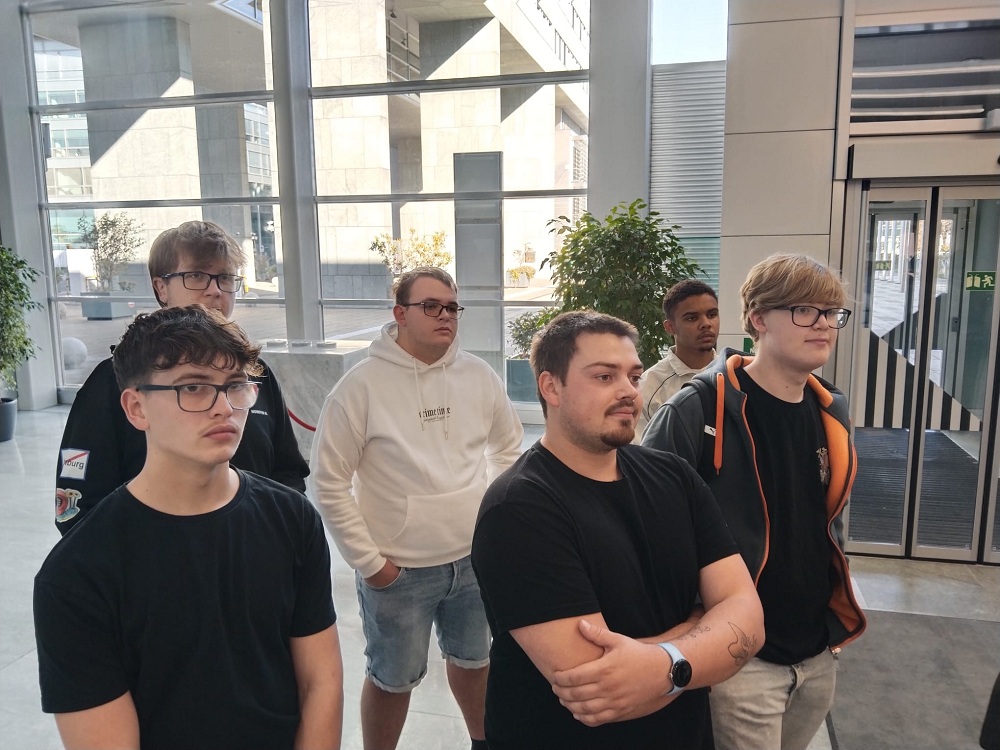 Sechs junge Männer stehen in einem modernen Gebäude mit großen Fenstern.